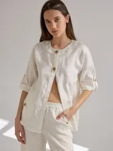 Camisa color crudo de lino, corte holgado, escote redondo, mangas 3/4 ajustables con botón y bolsillos delanteros con pespuntes a la vista.