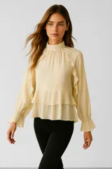 Blusa color crema de poliéster con cuello alto fruncido, mangas largas con puños elásticos y volados en el frente y en el ruedo. Corte holgado.
