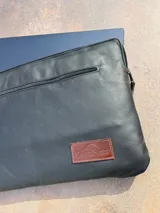 Funda negra de cuero para notebook, con cierre superior y bolsillo externo con cierre. Tiene un logo de la marca SierraMora en una etiqueta de cuero marrón cosida en la parte inferior derecha.