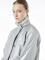 Campera plateada de poliéster con capucha ajustable y cintura con cordón ajustable interno. Cuenta con cierre frontal con botones a presión, bolsillos laterales con cierre y mangas 3/4 con puños elásticos.