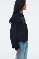 Blusa negra de manga larga con corte holgado y tejido texturizado. Presenta cuello corto con volados y abertura frontal con lazos para atar. Tiene canesú con inserciones de encaje y volados sobre los hombros, y puños largos con elástico y volado.