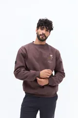 Buzo de algodón color marrón oscuro (ciruela/ciruela quemado) con cuello redondo, puños y ruedo acanalados. Presenta un pequeño bordado de motivos florales en el pecho.