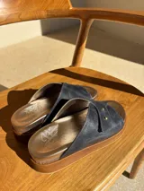 Sandalias de cuero color azul marino con plantilla dorada y suela marrón. Presentan una tira ancha cruzada con doble remache dorado.