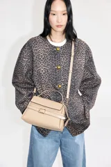 Cartera bandolera color visón, con correa ajustable y asa de mano. Presenta solapa con cierre magnético y trabilla decorativa con hebilla metálica.