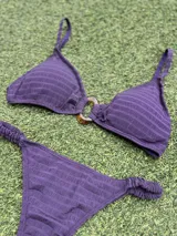 Conjunto de bikini color violeta confeccionado en tela texturada. El corpiño es de estilo top con una hebilla ovalada marrón en el centro y breteles finos. La bombacha es de corte colaless con detalle de frunces en los laterales.