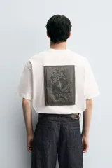 Camiseta blanca de corte holgado, cuello redondo y manga corta. Presenta un parche bordado en la espalda con diseño a contraste.