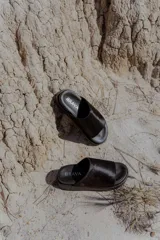 Sandalias planas tipo mule negras, con tira ancha superior de pelo y suela de caucho expandido. Llevan el logo BRAVA bordado en el lateral de la plantilla.