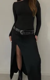 Vestido largo negro de mangas largas, con escote redondo y tajo lateral. Incluye cinturón de cuero negro con hebilla plateada y tachas.