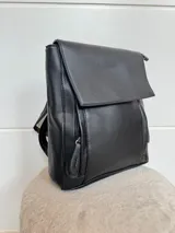Mochila color marrón de cuero sintético, con asa de mano y correas ajustables. Tiene un bolsillo frontal con cierre a presión y un bolsillo lateral con cierre.