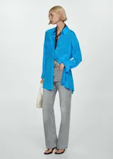 Camisa celeste de tejido satinado, con cuello camisero, cierre frontal asimétrico con botones, manga larga con puños abotonados, diseño ajustable con cuerda en la parte posterior y corte largo.