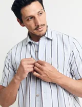 Camisa de manga corta con rayas verticales en tonos celeste, blanco, tostado y negro. Tiene cuello clásico, cierre con botones y bolsillo en el pecho.