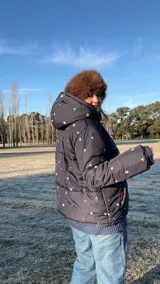 Campera puffer negra con estampado de flores y aves, capucha con borde de piel sintética marrón, bolsillos laterales y forro de algodón.