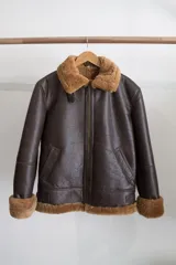 Campera gamulán marrón con cuello, puños y ruedo de piel sintética. Cierre frontal con cremallera y bolsillos laterales.