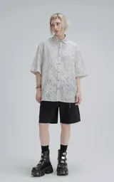 Camisa de manga corta color gris claro con cuello camisero y botones frontales. La tela presenta un bordado tipo broderie con motivos florales y geométricos.