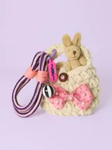 Cartera tejida color crema con asa de soga a rayas violetas y rosadas. Incluye un conejo de peluche beige en su interior y está decorada con un moño rosa con lunares blancos y un botón marrón.