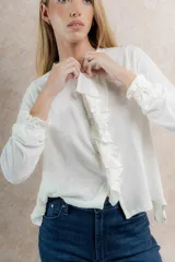 Blusa color crudo confeccionada en lino, con volados y botones en la delantera, cuello a la base y manga larga con elástico en los puños.