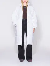 Plumífero blanco de corte recto y largo maxi, con cuello alto y cierre frontal con botones a presión. Incluye cinturón en la cintura.