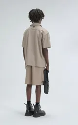 Bermuda estilo técnico color beige, confeccionada en microfibra liviana. Presenta cintura elástica con cinturón integrado de cinta negra con hebilla de plástico. Cuenta con múltiples bolsillos cargo, incluyendo uno lateral con panel de malla.