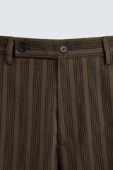 Pantalón marrón con finas rayas verticales negras, de corte amplio tipo balloon fit, con bolsillos laterales y traseros ocultos. Cierre frontal con cremallera y botonadura cruzada.