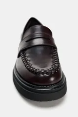 Zapato plano tipo mocasín color borgoña, con detalle de pasados en la parte delantera, copete subido y antifaz. Acabado en punta redondeada y suela track color negro.