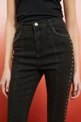 Pantalón de jean de tiro alto, color marrón oscuro con efecto lavado o desgastado, corte flare y tajo en el bajo. Presenta tachas doradas a lo largo de las costuras laterales.