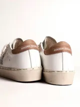 Zapatillas urbanas Golden Goose modelo Hi Star, de cuero blanco con detalles en gris y marrón. Presentan una estrella plateada con glitter en el lateral y suela de goma con inscripciones.