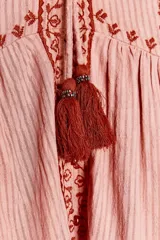Camisa de algodón color rosa con rayas verticales finas en rojo. Tiene cuello redondo con volantes, escote pico, mangas largas con puños y bordados combinados a contraste en rojo. Cierre frontal con botones y lazada con pompones.