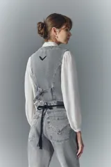 Chaleco de denim rígido lavado a gris claro, con botones metálicos grabados, bolsillos con solapa en el frente y tira de ajuste en la espalda. Presenta detalles de pasacintos en el escote.