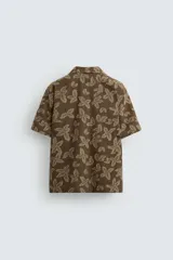 Camisa marrón con estampado floral, corte relaxed fit, cuello bowling, manga corta, bolsillo en el pecho, aberturas laterales y cierre con botones.