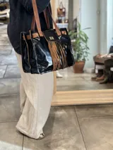 Bolso tipo tote de cuero negro brillante con doble asa de cuero marrón y flecos de cuero marrón en la parte frontal.