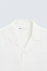 Camisa de calce relajado confeccionada en tejido fluido de mezcla de viscosa, color blanco. Presenta cuello bowling, manga corta, aberturas laterales en el bajo y cierre frontal con botones al tono.