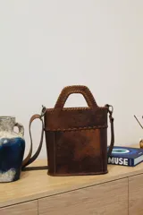 Bolso matero de cuero color marrón con manija superior trenzada y correa larga desmontable.