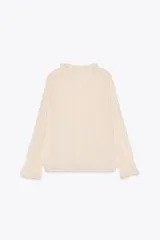 Blusa color crema semitransparente con cuello redondo con volantes y cordón. Manga larga acabada en elástico y volante. Cierre frontal con botones.