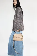 Cartera bandolera color visón, con correa ajustable y asa de mano. Presenta solapa con cierre magnético y trabilla decorativa con hebilla metálica.