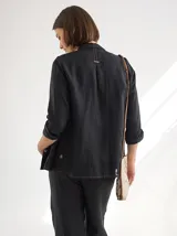 Camisa negra de lino con corte femenino, cuello clásico, cierre frontal con botones y mangas 3/4 con puños enrollados. Presenta detalles de frunces verticales en el frente y bolsillos de parche a la altura de la cadera.
