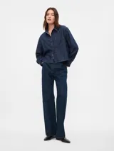 Camisa corta de jean azul oscuro, con cuello clásico, cierre frontal con botones, manga larga y bolsillo de parche en el pecho.