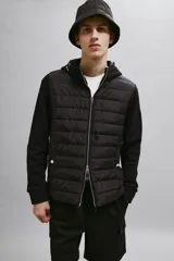 Campera negra híbrida con cuerpo acolchado y mangas de punto. Tiene capucha con cordones ajustables, cierre frontal, bolsillos laterales con botón a presión y puños acanalados.