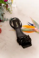 Sandalia negra de charol con taco cuadrado, tira fina al tobillo con hebilla y moño trenzado en el empeine.