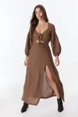 Vestido color beige de lino, con escote en V, mangas largas abullonadas y lazos frontales. Tiene corte a la cintura y falda larga con tajo frontal.
