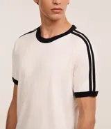 Remera de algodón de corte regular, color blanco hueso, con cuello redondo y ribete negro. Presenta un diseño de doble raya negra en las mangas y ribetes negros en el cuello y los puños de las mangas.
