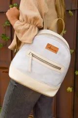 Mochila pequeña de cuero color gris claro y beige, con cierre superior y bolsillo frontal con cierre. Tiene asa de mano y correas ajustables para los hombros.