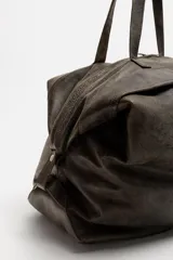 Bolso de viaje marrón oscuro fabricado en piel con acabado craquelado. Tiene un compartimento principal con cierre de cremallera, un bolsillo plano interior con cremallera y dos bolsillos frontales con botón a presión. Cuenta con asas de mano de piel y asa de hombro removible y ajustable.