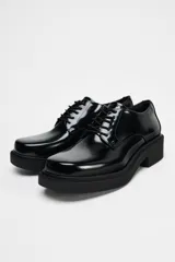 Zapato tipo mocasín negro acharolado con punta redonda y cierre de cordones. Suela de 4 cm de altura.
