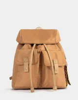 Mochila color marrón claro de tela deportiva con solapa, correas regulables, manija superior, cierre con broche a presión y tira de ajuste. Tiene doble bolsillo frontal con broche a presión y charm lateral en forma de cartera.