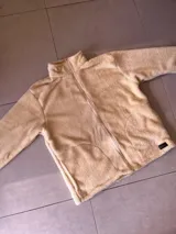 Buzo de poliéster color crema, con textura de sherpa, cuello alto y cierre frontal con cremallera.