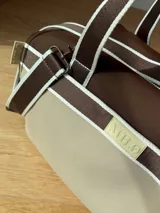 Bolso tipo tote de neopreno color negro y marrón, con cierre superior, bolsillo frontal marrón con logo rojo, bolsillo interno y externo con cierre, doble asa de mano marrón con detalles blancos y opción de correa larga.