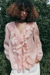 Camisa color rosa pálido confeccionada en tejido ligero con lyocell. Presenta escote en V, cierre frontal con botones, mangas largas abullonadas con puños elásticos y detalle de volantes en el frente.