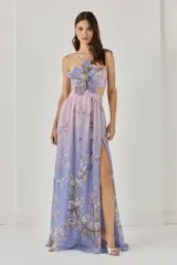 Vestido largo de gasa con estampado floral en tonos lila y rosa, con una flor bordada en 3D en el busto, corte en la cintura y falda amplia con abertura lateral. Cuenta con botones y cierre trasero con cremallera invisible.