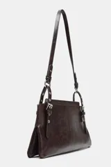 Bolso de hombro de formato alargado, color marrón oscuro con acabado ligeramente desgastado. El asa de hombro es delgada y ajustable mediante hebillas metálicas. Presenta detalles de tachas o remaches metálicos en los puntos de unión del asa.
