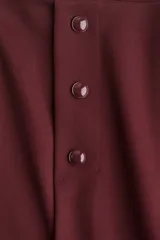 Short pollera mini de tejido de punto pesado, color borgoña, de tiro alto con elástico oculto en la cintura. Presenta pliegues y una hilera de botones decorativos en el frente.